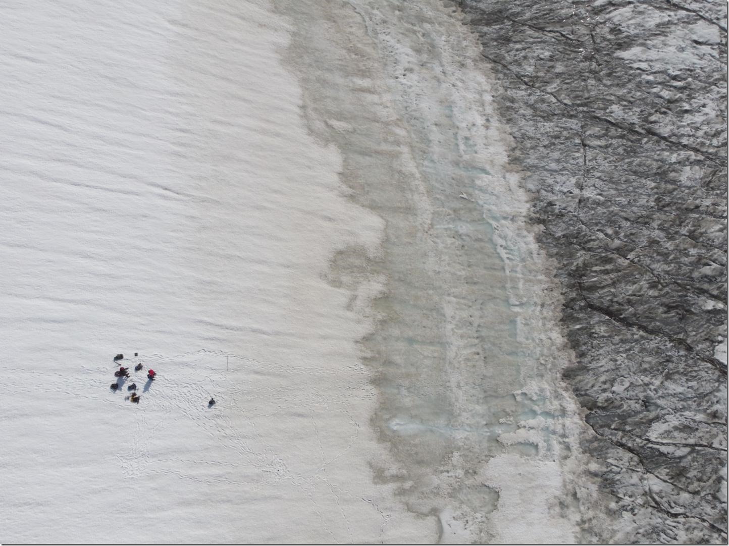 This photo shows the change in surface darkness as the cold dry white snow (left side of of image) starts melting to grey (centre of the image), to reveal the dark ice underneath (right side of image). The dots on the left are people to give a sense of the scale. Credit Laura Halbach