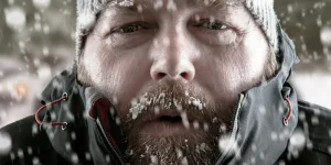 Freezing cold man standing in a snow storm blizzard trying to keep warm. Wearing a beanie hat and winter coat with frost and ice on his beard and eyebrows staring at the camera.